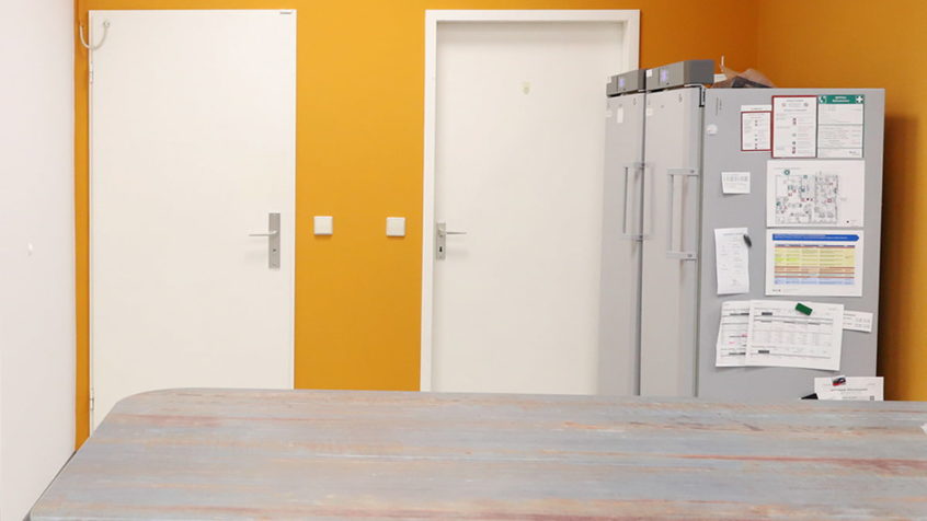 Äskulap Apotheke Riesa-Weida Backoffice mit orangefarbener Wand, zwei Kühlschränken, zwei weißen Türen und einer Tischplatte im Vordergrund
