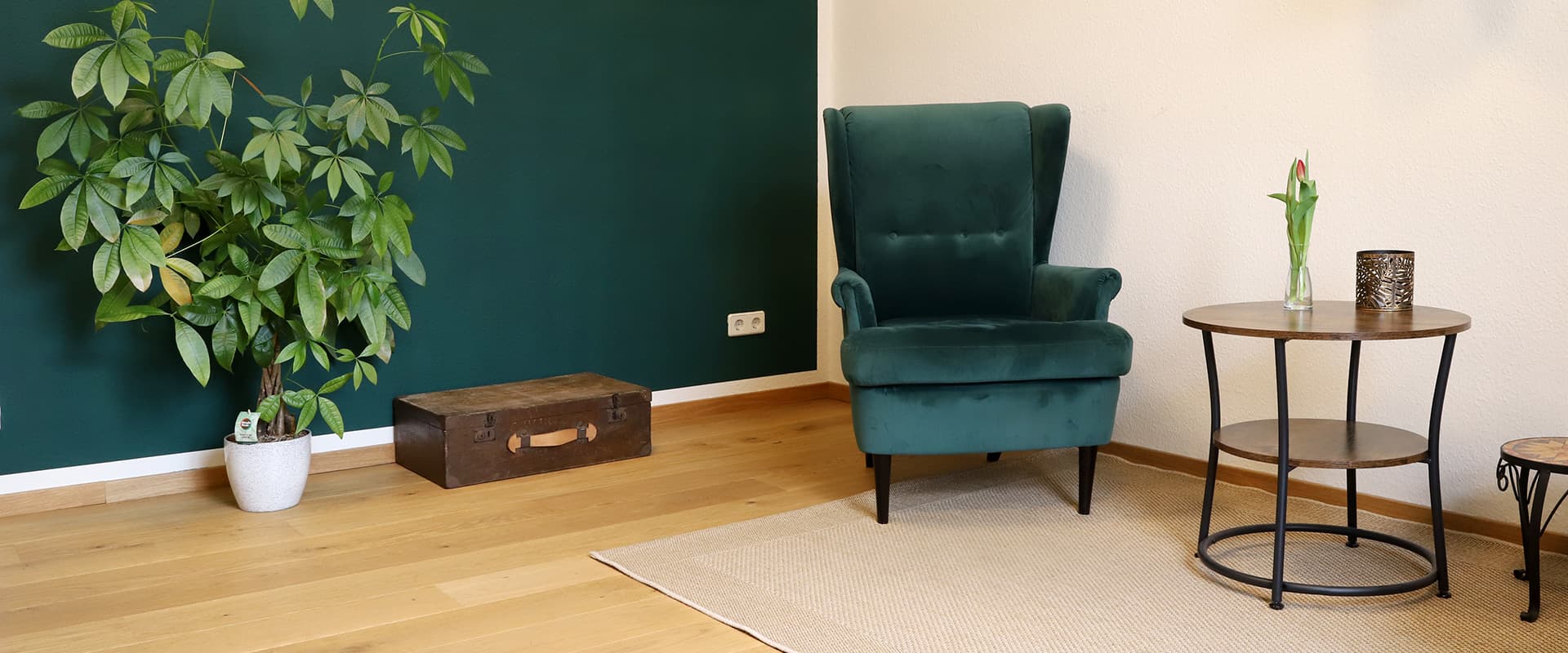 Referenz Praxis für Psychotherapie Peterhänsel Leipzig grüner Sessel in Samtoptik auf einem Teppich, kleiner runder Tisch, einen Koffer sowie eine Pflanze vor einer petrolfarben gestrichenen Wand