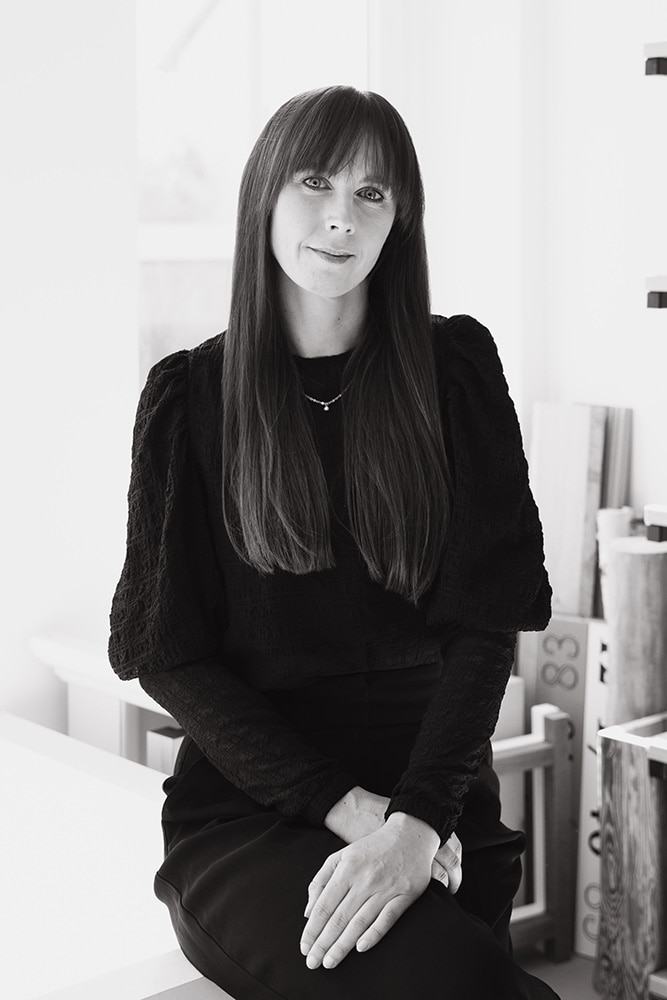 mediraum design Schwarz-Weiß-Bild - Elisa Borchert trägt ein dunkles Oberteil, eine dunkle Hose, eine Halskette & blickt leicht lächelnd in die Kamera