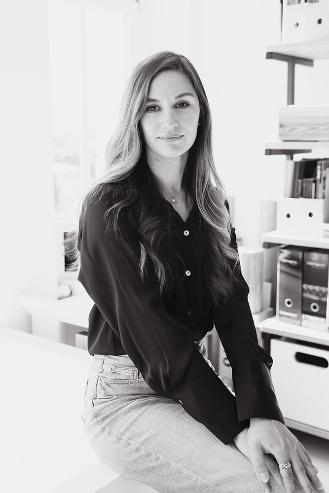 mediraum design Schwarz-Weiß-Bild - Karyna Stopicheva trägt ein dunkles Oberteil, eine helle Jeans, eine Halskette & blickt leicht lächelnd in die Kamera