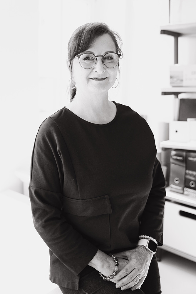 mediraum design Schwarz-Weiß-Bild - Kerstin Hornig trägt ein dunkles Oberteil, eine Brille & Schmuck an beiden Händen & blickt leicht lächelnd in die Kamera