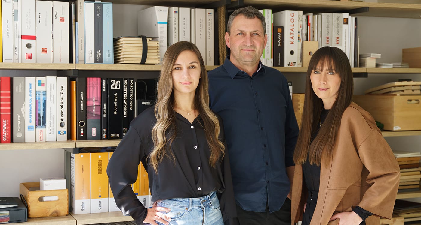 mediraum design Teamfoto Karyna Stopicheva, Hagen Brockhaus & Elisa Borchert stehen leicht lächelnd vor einem Regal mit vielen Büchern & Holzkisten